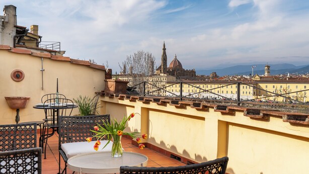 Eine Terrasse mit Sitzgelegenheiten im Freien, eine Vase mit Blumen und Blick auf eine Stadtlandschaft mit historischen Gebäuden.
