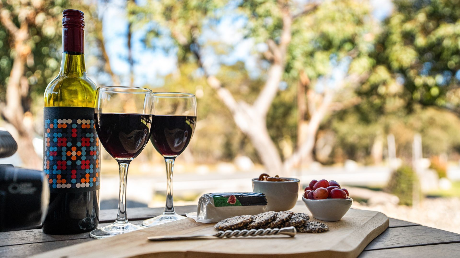 A bottle of wine, two wine glasses, a cheese, a bowl of grapes, and crackers on a wooden table outdoors.