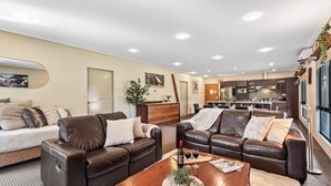 A modern living room with leather sofas, a wooden coffee table, and a kitchen in the background. - Kickenback Lodge - 3BR Lodge l Gym l Indoor Pool l Tennis l WiFi l Air-Con (Crackenback)