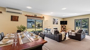 A dining area with a wooden table set for four, a sofa, and a television in a living room with a view of the outdoors.
