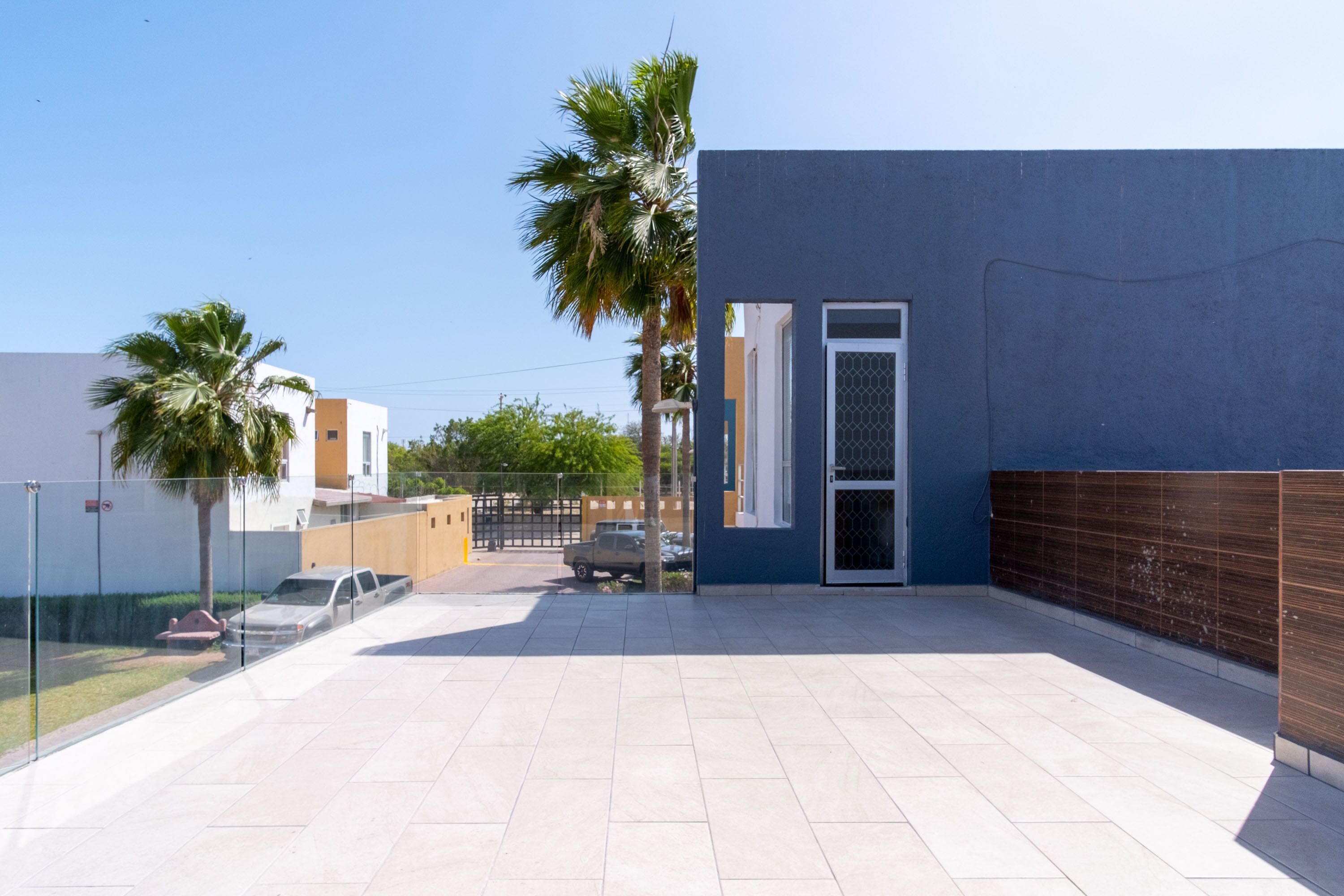 A modern building with a blue exterior, a gated entrance, and a paved patio area with palm trees.
