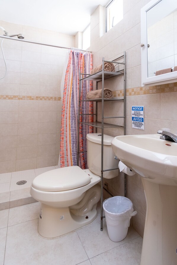 Un baño con inodoro, lavamanos y zona de ducha. Hay un cartel en la pared y un cubo de basura cerca del lavamanos.
