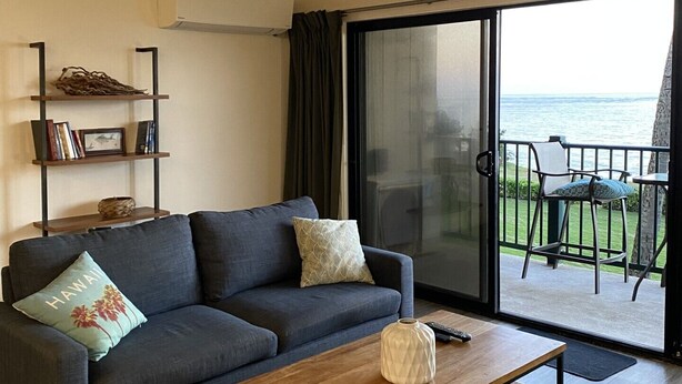 Sala de estar com sofá, mesa de centro, estante de livros e vista para o mar através de uma porta de vidro deslizante.