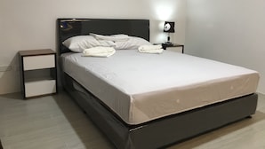 A neatly made bed with white linens and a folded towel, a bedside table with a lamp, and a nightstand with a power outlet. - Mayumi Lodge, Perfect for couples. Cozy and Nice! Fast Internet (Puerto Princesa)