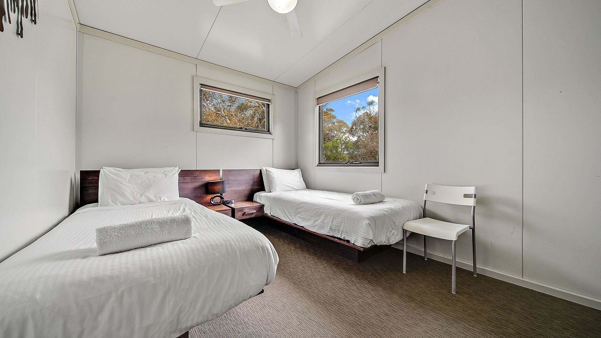 Habitación de hotel con dos camas, una mesita, una silla y grandes ventanales con vistas a árboles.
