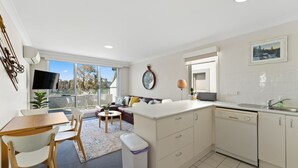 Una cocina moderna con armarios blancos, un área de comedor con mesa y sillas, un sofá y un ventanal con vistas a árboles.