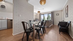 Une cuisine moderne avec des murs en briques blanches, un coin repas avec une table en verre et des chaises noires, et une grande fenêtre donnant sur l’extérieur.