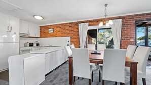 A modern kitchen with white cabinetry, a built-in oven, and a microwave. There is a dining area with a wooden table and white chairs.