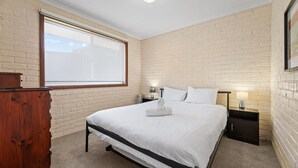 A bedroom with a large window, a bed with white linens, a wooden dresser, and a nightstand with a lamp.