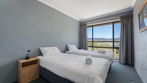 A hotel room with two beds, a wooden nightstand, a desk, and a large window offering a scenic view.