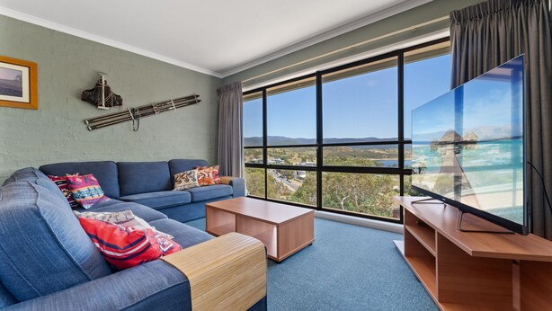 A modern living room with a large window offering a view of a tropical beach and overwater bungalows.