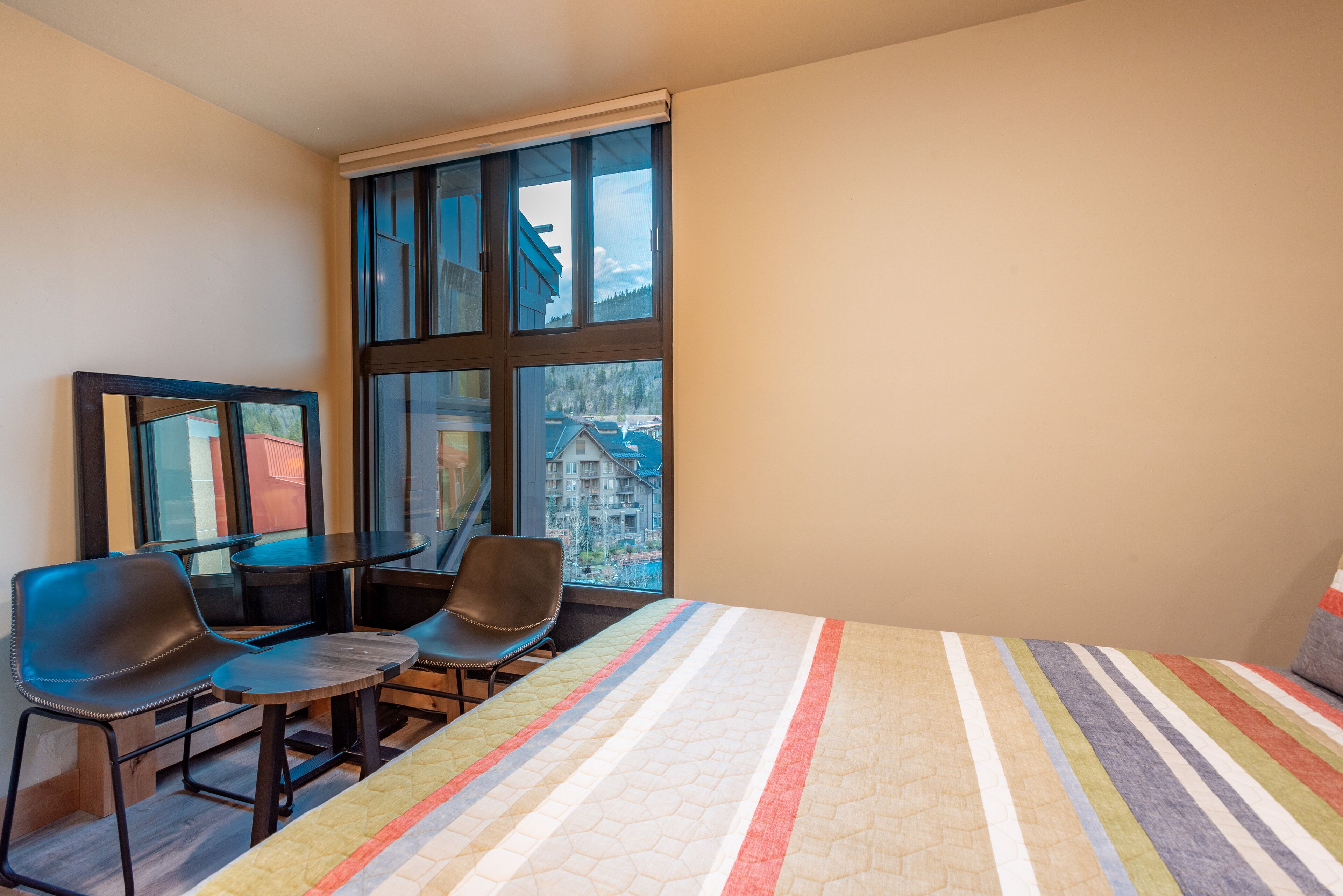 A bedroom with a large window, a bed with a striped quilt, two chairs, and a small table.