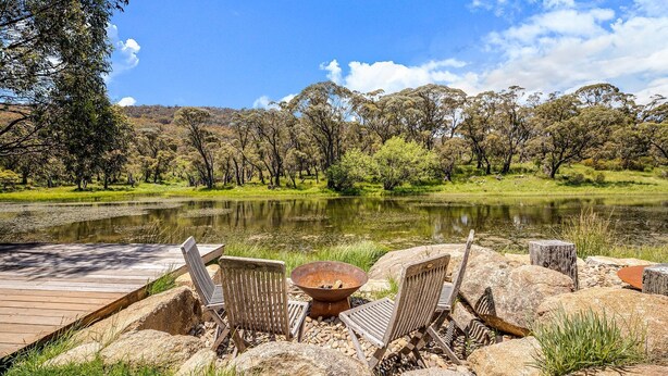 A lakeside seating area with wooden chairs and a fire pit, surrounded by rocks and trees.