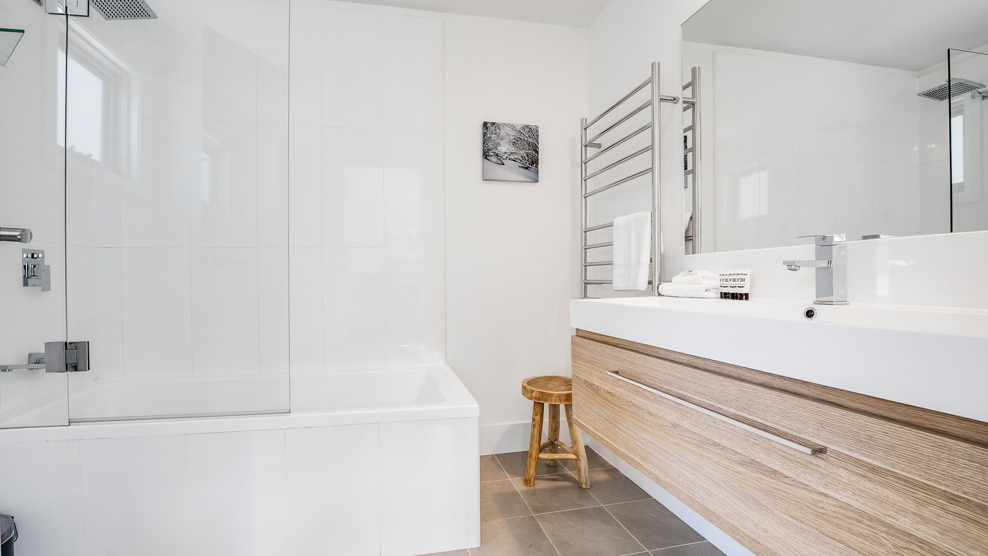 Un bagno moderno con cabina doccia in vetro, uno sgabello in legno, un mobile lavabo bianco con specchio e un quadro appeso alla parete.