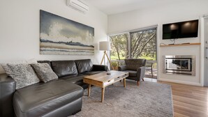 A modern living room with a leather sofa, a wooden coffee table, a grey armchair, a large painting, a floor lamp, a TV, and a built-in oven.