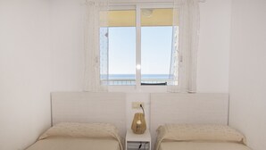 Ein Zimmer mit zwei Betten, einem kleinen Tisch mit Lampe, einem Fenster mit Vorhängen und Meerblick.
