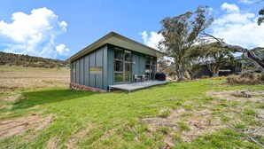A modern, single-story building with a flat roof and large windows, situated in a grassy field with a view of a hilly landscape. - Ecocrackenback Luxe Chalet 19 (CRACKENBACK)