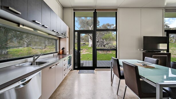 A modern kitchen with a large window, a dining area with a glass table, and a flat-screen TV. - Ecocrackenback Luxe Chalet 19 (CRACKENBACK)