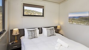 A neatly made bed with white linens and patterned pillows, a bedside table with a lamp, and a window with a view of a hilly landscape. - Ecocrackenback Luxe Chalet 19 (CRACKENBACK)