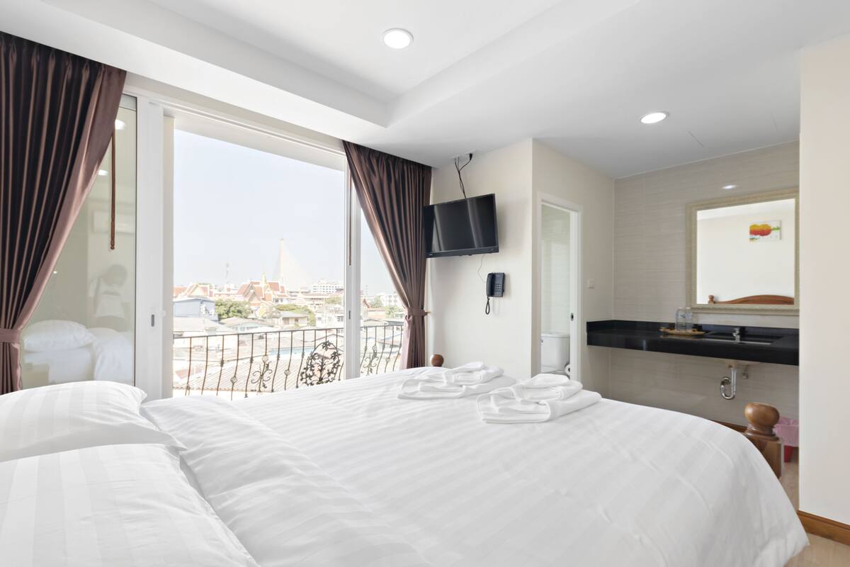A hotel room with a large bed, a TV mounted on the wall, a bathroom with a mirror, and a balcony with a view of the city.