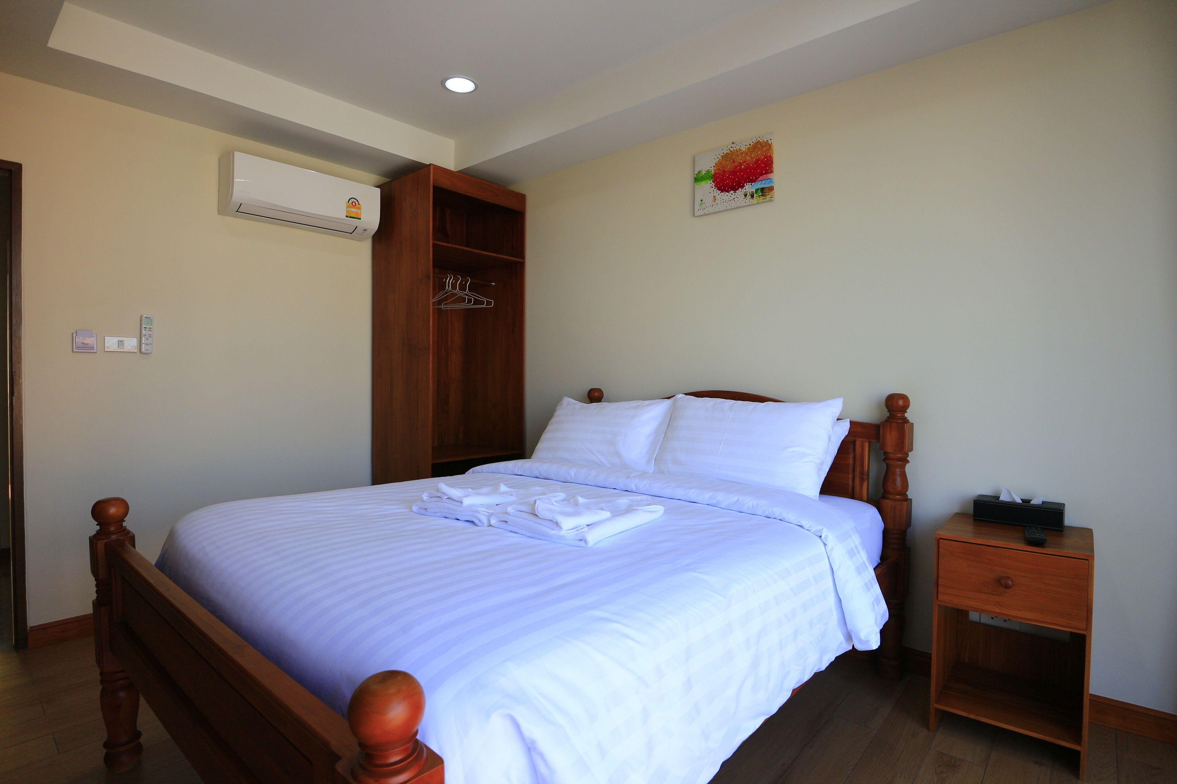 A neatly made bed with white linens and a wooden headboard, a wooden nightstand with a tissue box, and a wall-mounted air conditioner.