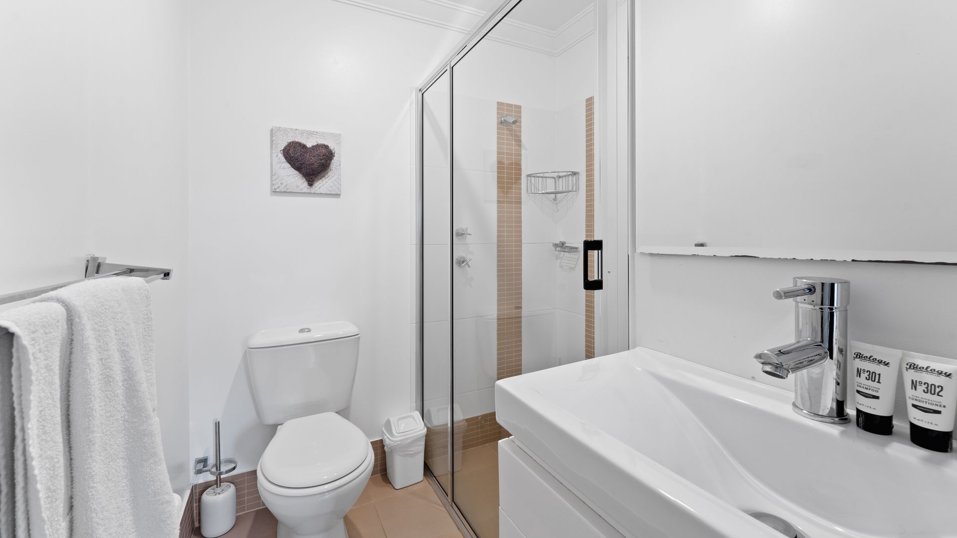A modern bathroom with a toilet, shower, and sink. There is a heart-shaped artwork on the wall and toiletries on the sink.