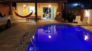 Una piscina con borde iluminado en azul, una hamaca y una casa con techo de tejas al fondo.