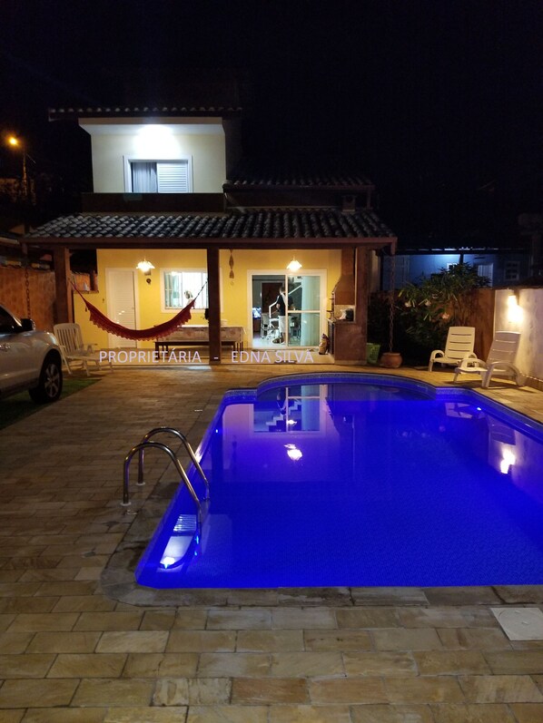 Una piscina con borde iluminado en azul, una hamaca y una casa con techo de tejas al fondo.