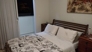 Un dormitorio con una cama de madera, ropa de cama blanca, una colcha con motivos de periódico, una mesita de noche, una lámpara y una ventana con cortinas.