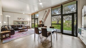 Una sala de estar moderna con sofá, mesa de centro y comedor. Amplias ventanas con vista a árboles y una chimenea.