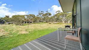 A balcony with a grill and seating, overlooking a grassy area with trees and a building in the background. - Ecocrackenback 16 'Sustainable chalet close to the slopes.' (CRACKENBACK)