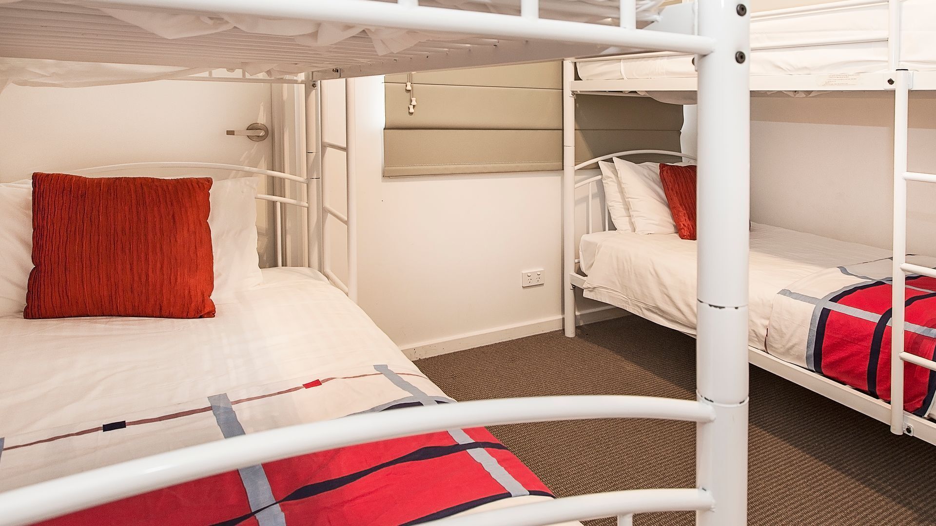 Une chambre avec un lit superposé à la structure métallique blanche, un oreiller rouge et une couverture rouge et noire.