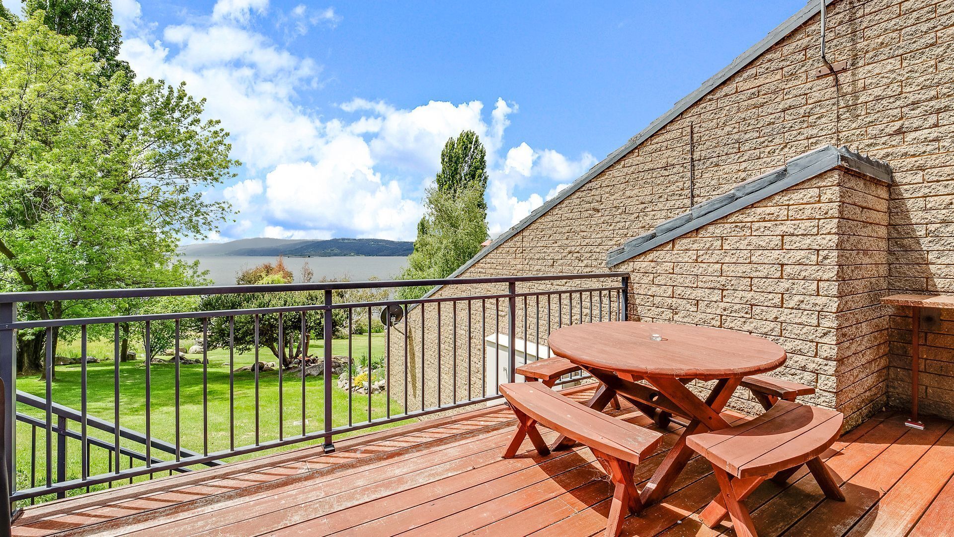 Une terrasse en bois, dotée de tables de pique-nique et d’une rambarde en métal, qui offre une vue sur une zone herbeuse et une étendue d’eau.