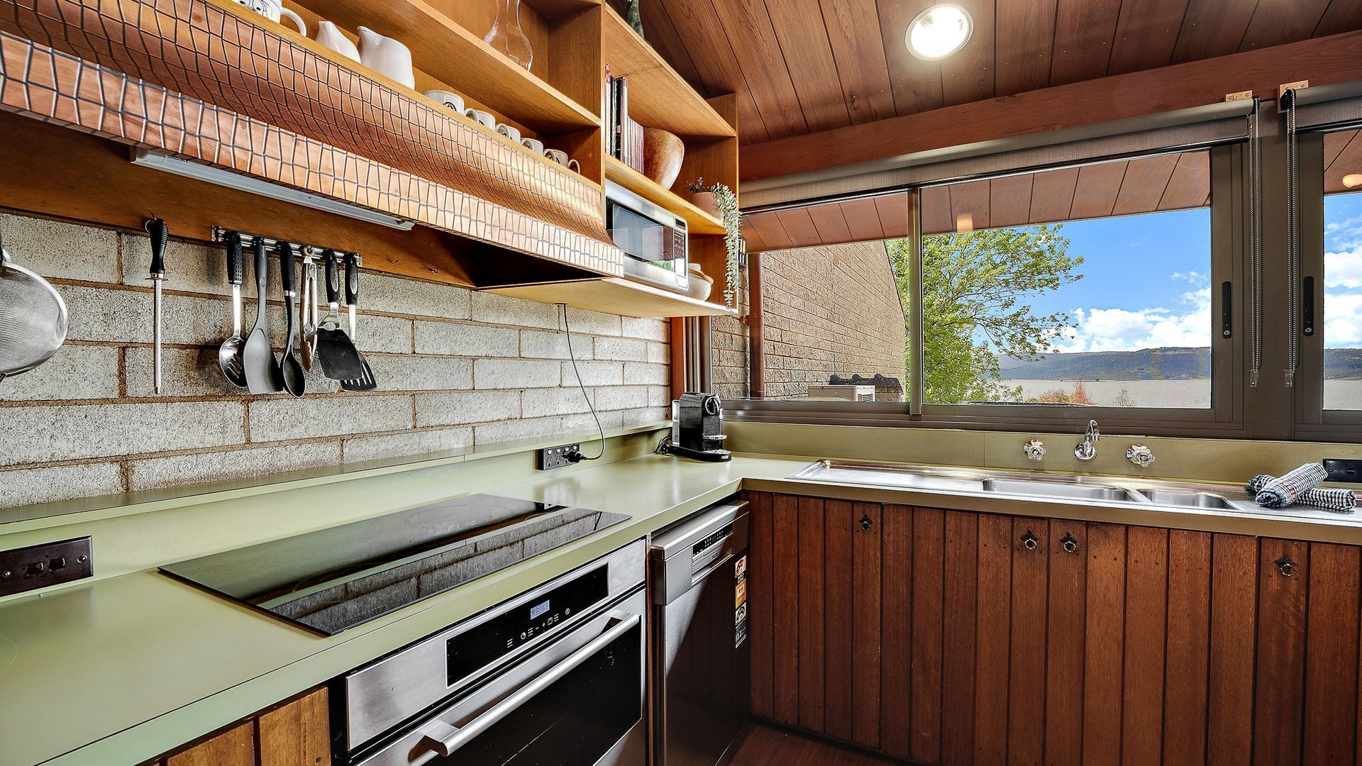 A kitchen with a brick backsplash, wooden cabinets, a stainless steel sink, and a window overlooking a scenic landscape.