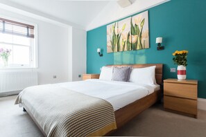 Une chambre à coucher avec un lit, des tables de chevet, une fenêtre avec des stores, un mur d’accent de couleur turquoise et un tableau.