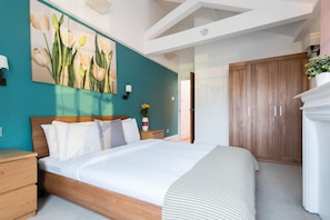 A bedroom with a bed, wooden headboard, teal walls, a large floral painting, a nightstand with a vase, and a wardrobe.