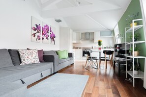 Un salon moderne avec un canapé gris, une table basse et un coin repas comprenant une table et des chaises. Au fond, on aperçoit une cuisine avec des meubles blancs et une fenêtre.