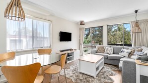 A modern living room with a glass dining table, wooden chairs, a grey sofa, and a flat-screen TV mounted on the wall.
