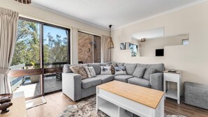 A modern living room with a grey sectional sofa, a wooden coffee table, and a large window offering a view of trees.