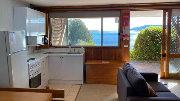 A compact kitchen with a view of the sea, a sofa, and a fire blanket sign. - Seeblick 3 - 1BR Lakefront Apartment l Central Location l Off Street Parking (JINDABYNE)