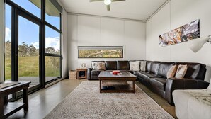 A spacious living room with a large window, a leather sofa, a coffee table, and a rug. - Ecocrackenback 7 'Sustainable chalet close to the slopes.' (Crackenback)