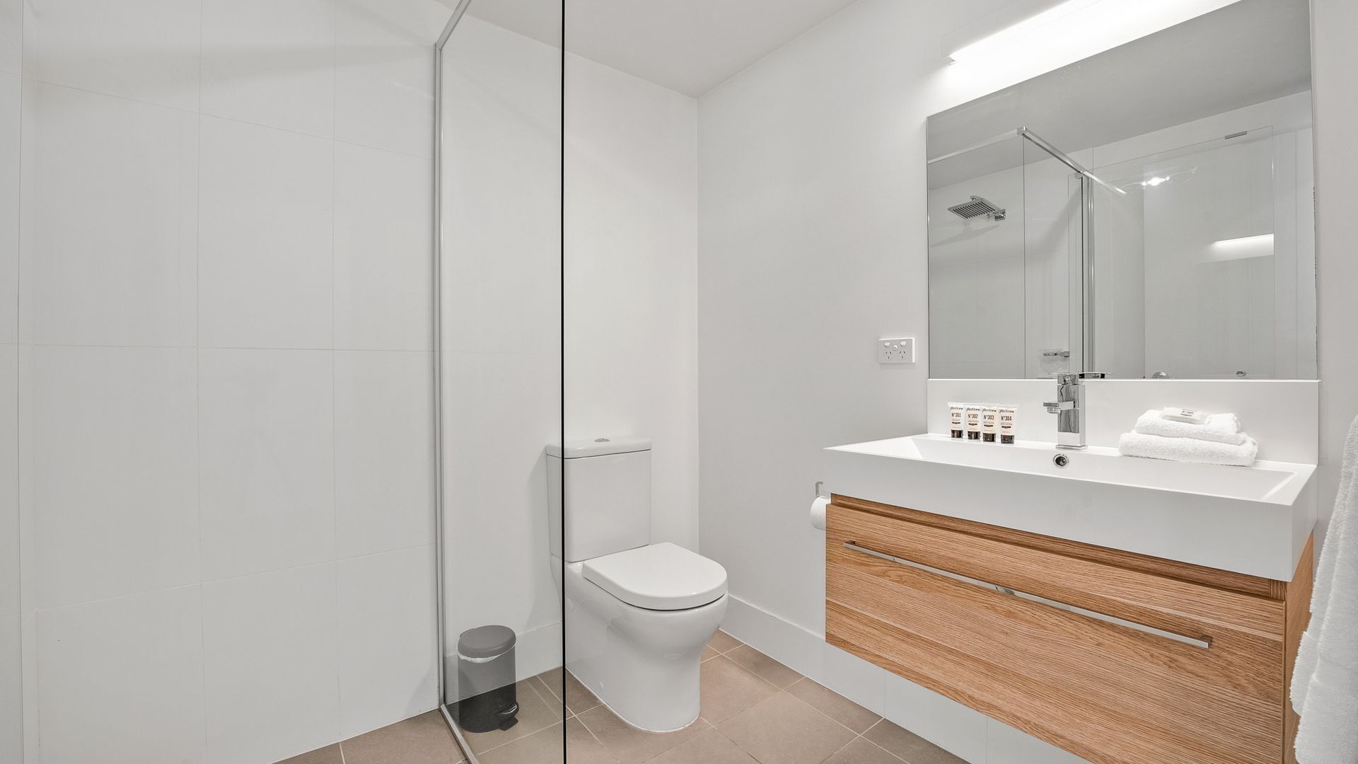 A modern bathroom with a glass shower enclosure, a toilet, a sink with toiletries, and a wooden vanity.