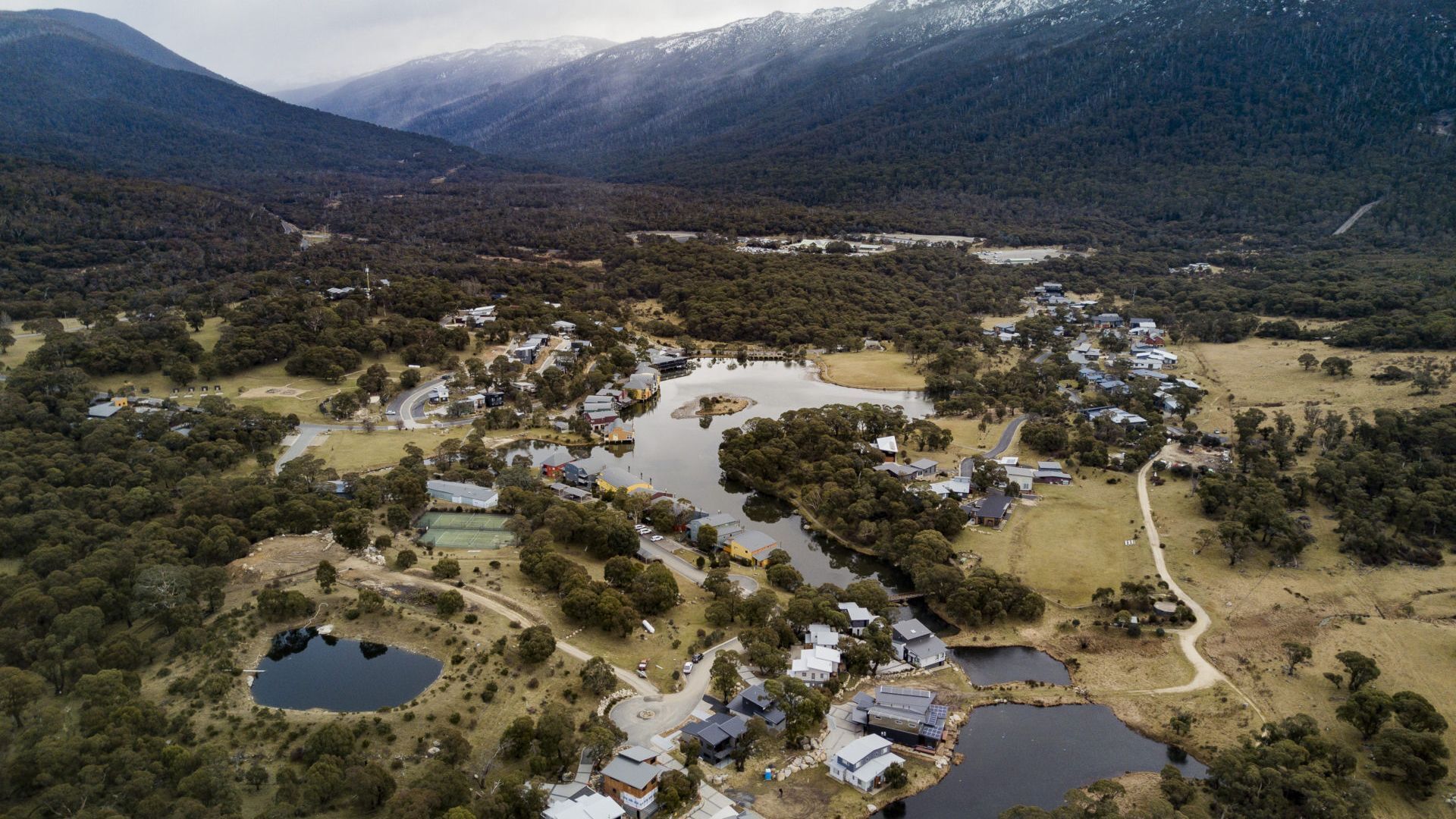 An aerial view of a residential area with houses, a lake, and surrounding forested hills.