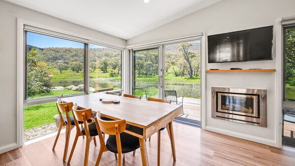 Un espace repas comprenant une grande table et des chaises, une cheminée et de grandes fenêtres donnant sur un lac et des arbres.