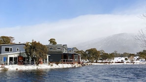 Une propriété située au bord de l’eau, avec des maisons modernes, un quai et un paysage enneigé, agrémenté d’arbres et d’une montagne en arrière-plan.