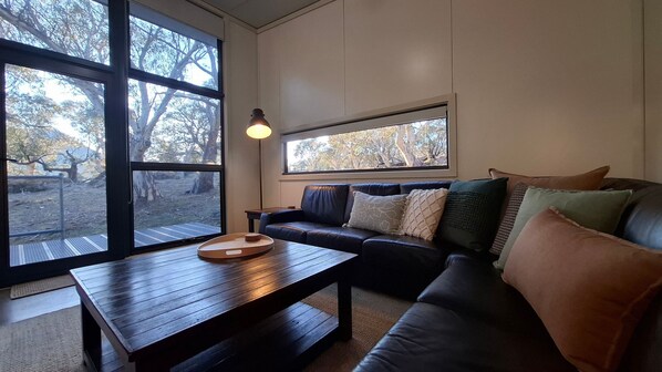 A living room with a dark leather sofa, a wooden coffee table, and a large window providing a view of trees.