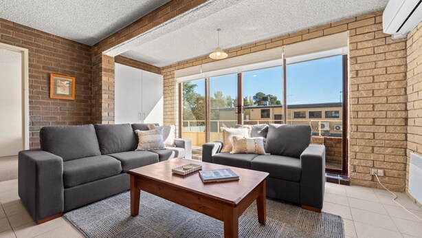 A modern living room with two grey sofas, a wooden coffee table, and large windows offering a view of buildings. - Central Park 4 - 3BR Ground Floor Apartment l Central Location (Jindabyne)