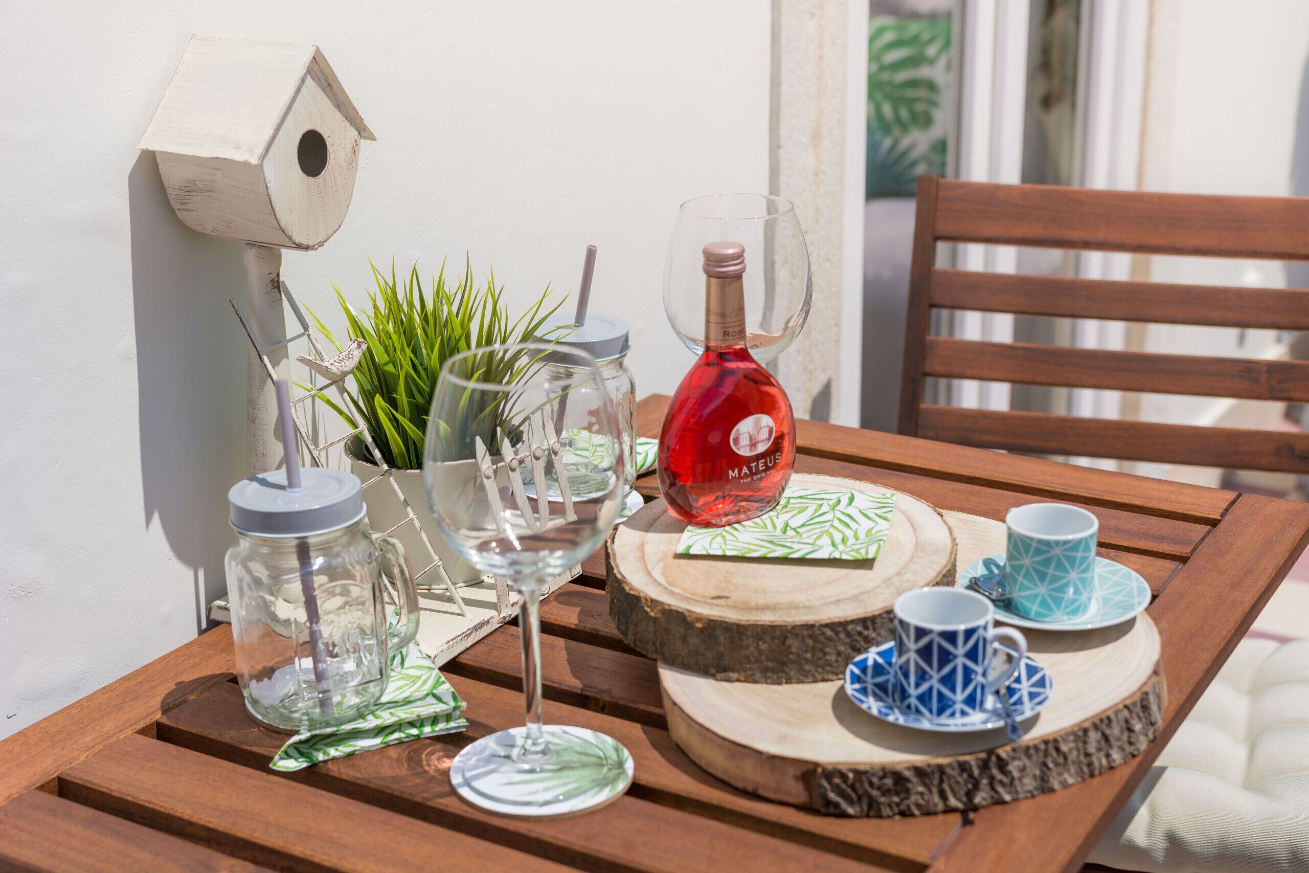 A wooden table with a birdhouse, a potted plant, a wine bottle, and a set of blue and white cups and saucers.