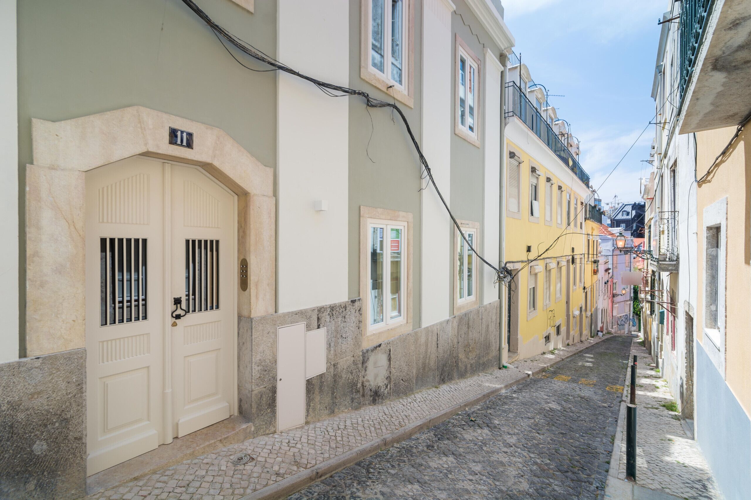 A narrow cobblestone street flanked by buildings with traditional architecture, including a white door with barred windows and a yellow building with a balcony.