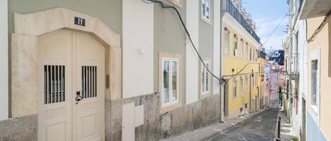 A narrow cobblestone street flanked by buildings with traditional architecture, including a white door with barred windows and a yellow building with a balcony.